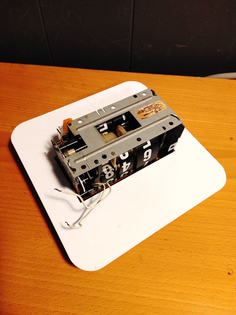 Underside/back view of a 3D printed faceplate with an analog clock timepiece sitting inside it. In this image you can see more of the clock mechanism.